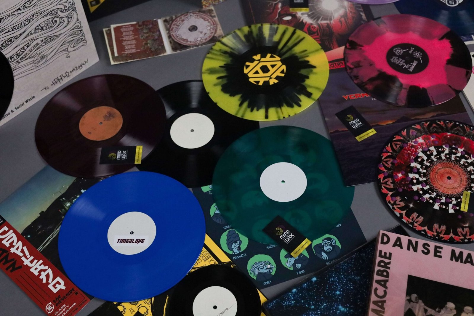 Collection of vinyl records pressed by Mind The Wax, featuring black, colored, splatter, smash vinyl, and a box set displayed on the floor.