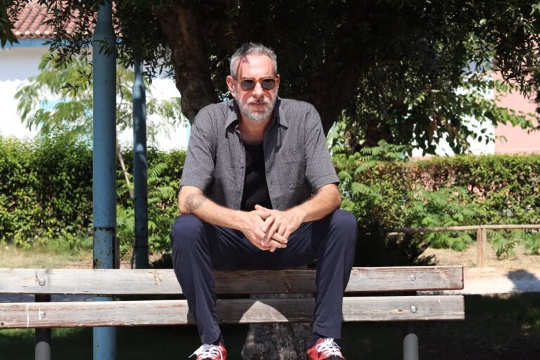 Blend Mishkin sitting on a bench outdoors wearing sunglasses and a shirt